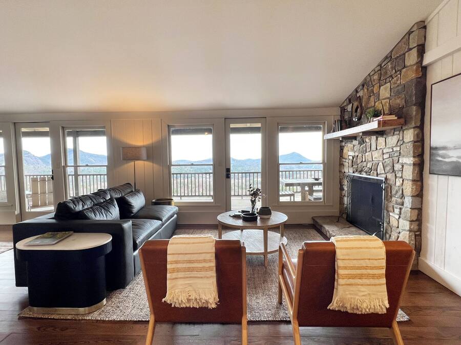 A living room with a sofa, chairs, a coffee table, and a fireplace. There are large windows with a view of mountains.