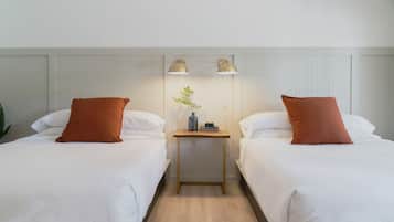 A hotel room with two beds, each with white bedding and a brown pillow. There is a small wooden nightstand between the beds with two lamps and a vase with a plant.