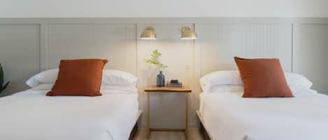 Habitación de hotel con dos camas, cada una con ropa blanca de cama y almohada marrón. Entre las camas hay una mesita de noche de madera con dos lámparas y un florero con una planta.