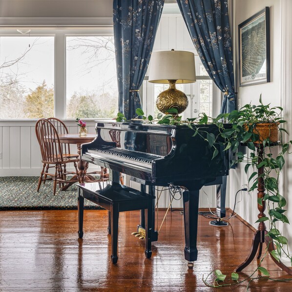 A grand piano in a room with large windows, a table with chairs, and a potted plant.