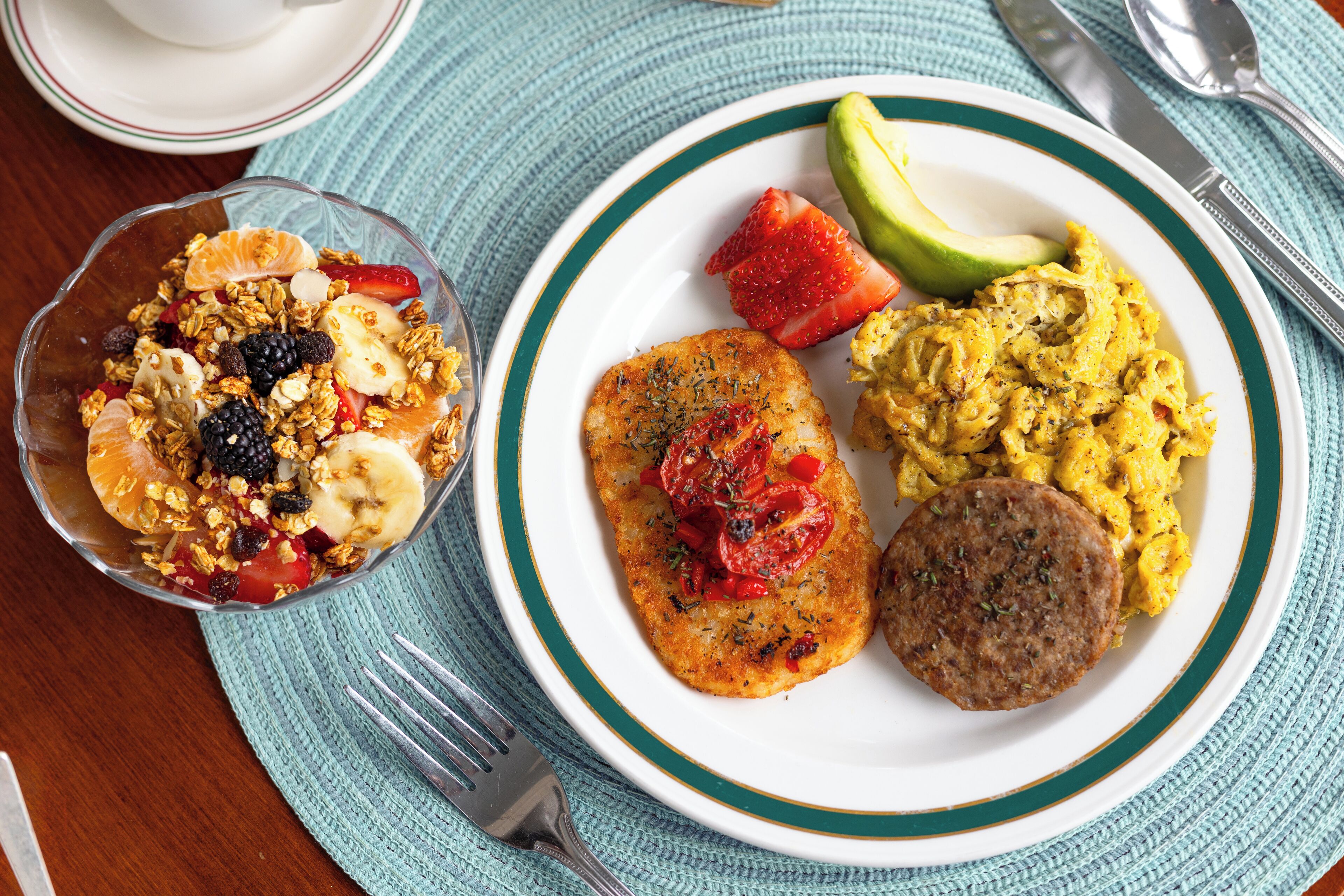 A breakfast plate with scrambled eggs, a patty, and a bowl of fruit and granola.