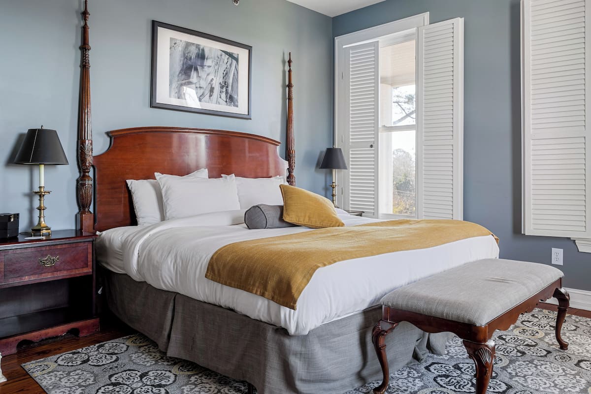 A bedroom with a large bed, a nightstand, a bench, and a window with shutters.