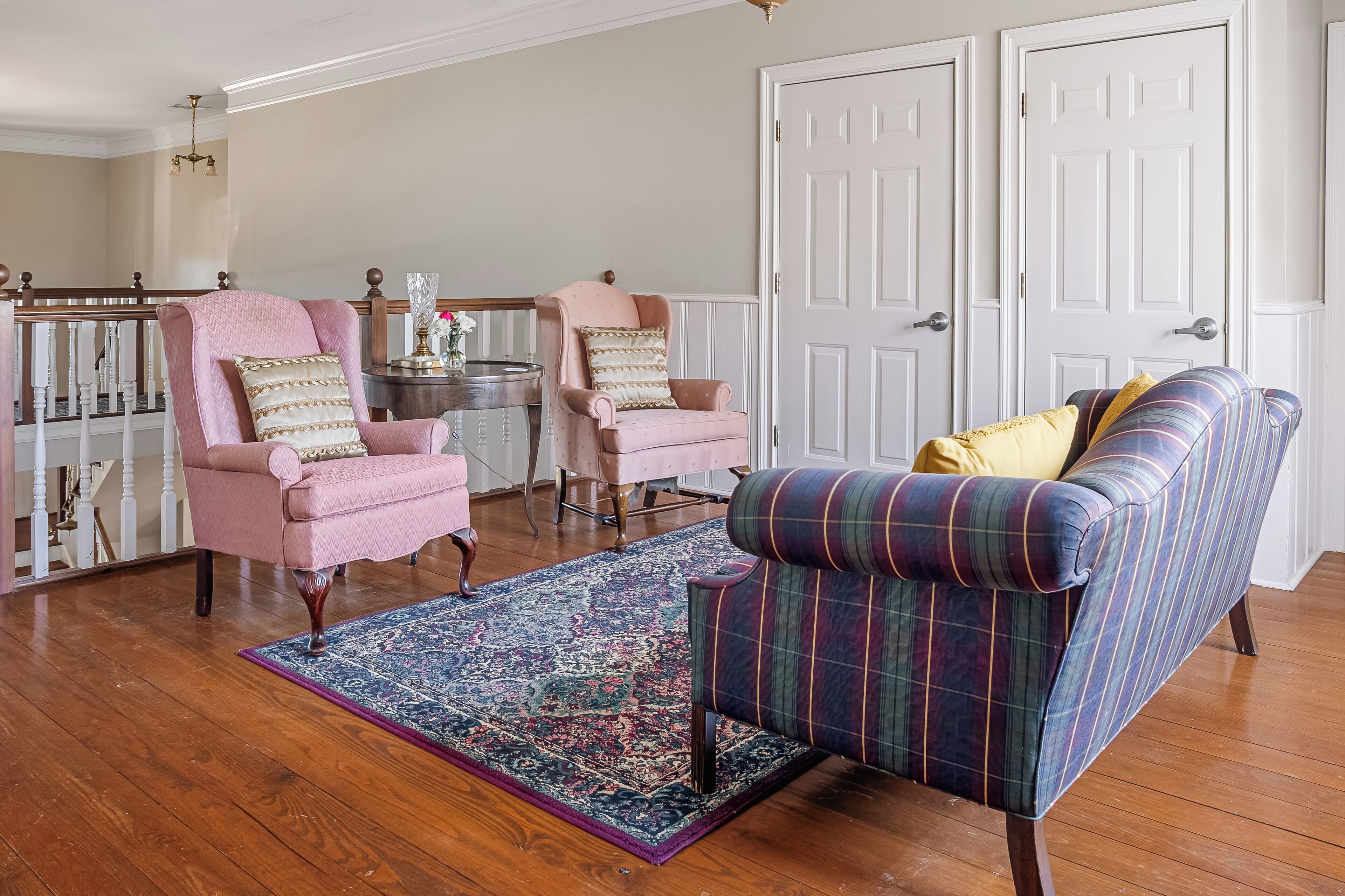 A living room with two pink armchairs, a plaid sofa, a patterned rug, and a wooden staircase.