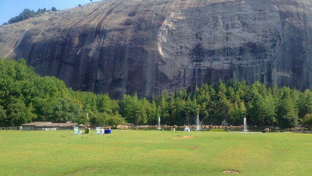 A large rock formation with a carved relief, surrounded by a grassy area with trees and a small building. - Stone Mountain Manor (Stone Mountain)