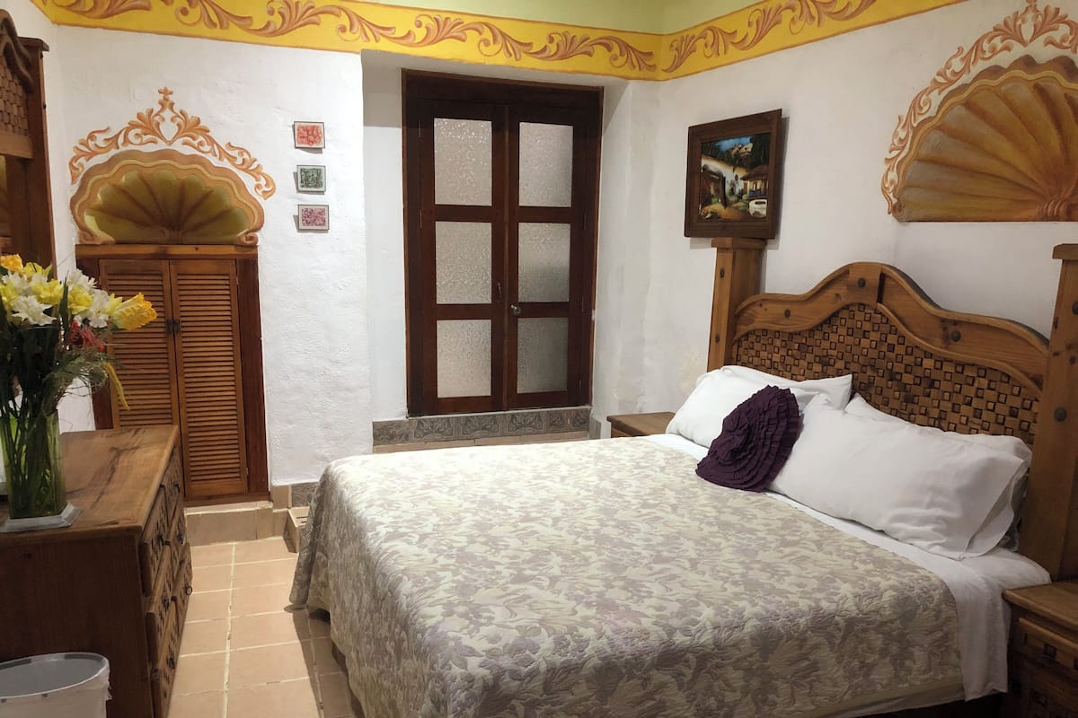 Ein Schlafzimmer mit einem Bett, einem Holzkopfende, einem Nachttisch, einer Kommode, einem Spiegel, einer Tür mit Glasfeldern und einer Wand mit dekorativen Elementen.