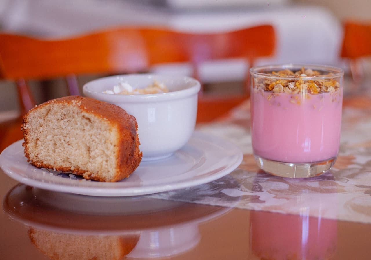 A slice of cake on a plate, a bowl of cereal, and a glass of pink smoothie with granola on a table.