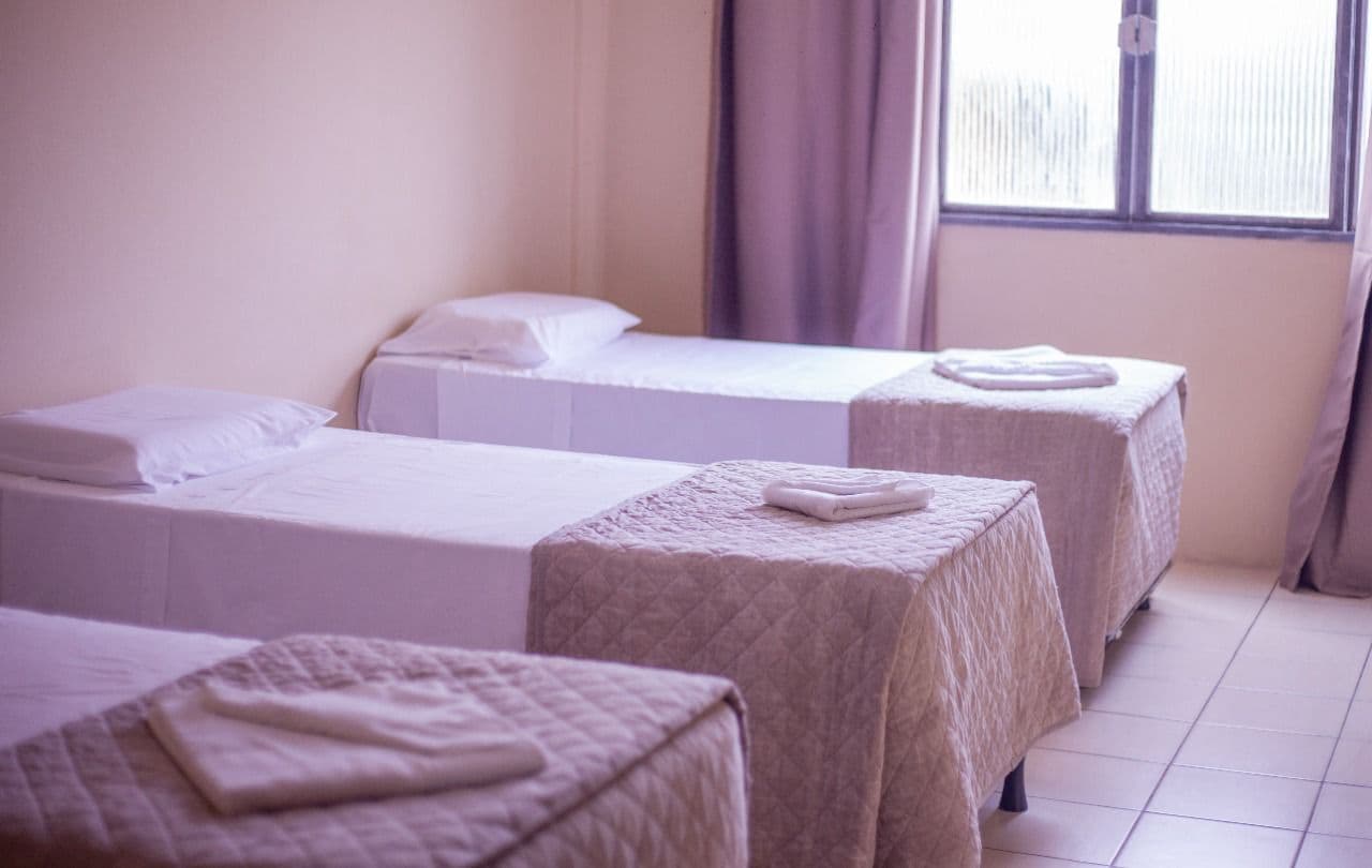 A hotel room with three beds, each with a white bedspread and pillows. The room has a window with curtains, and the floor is tiled.