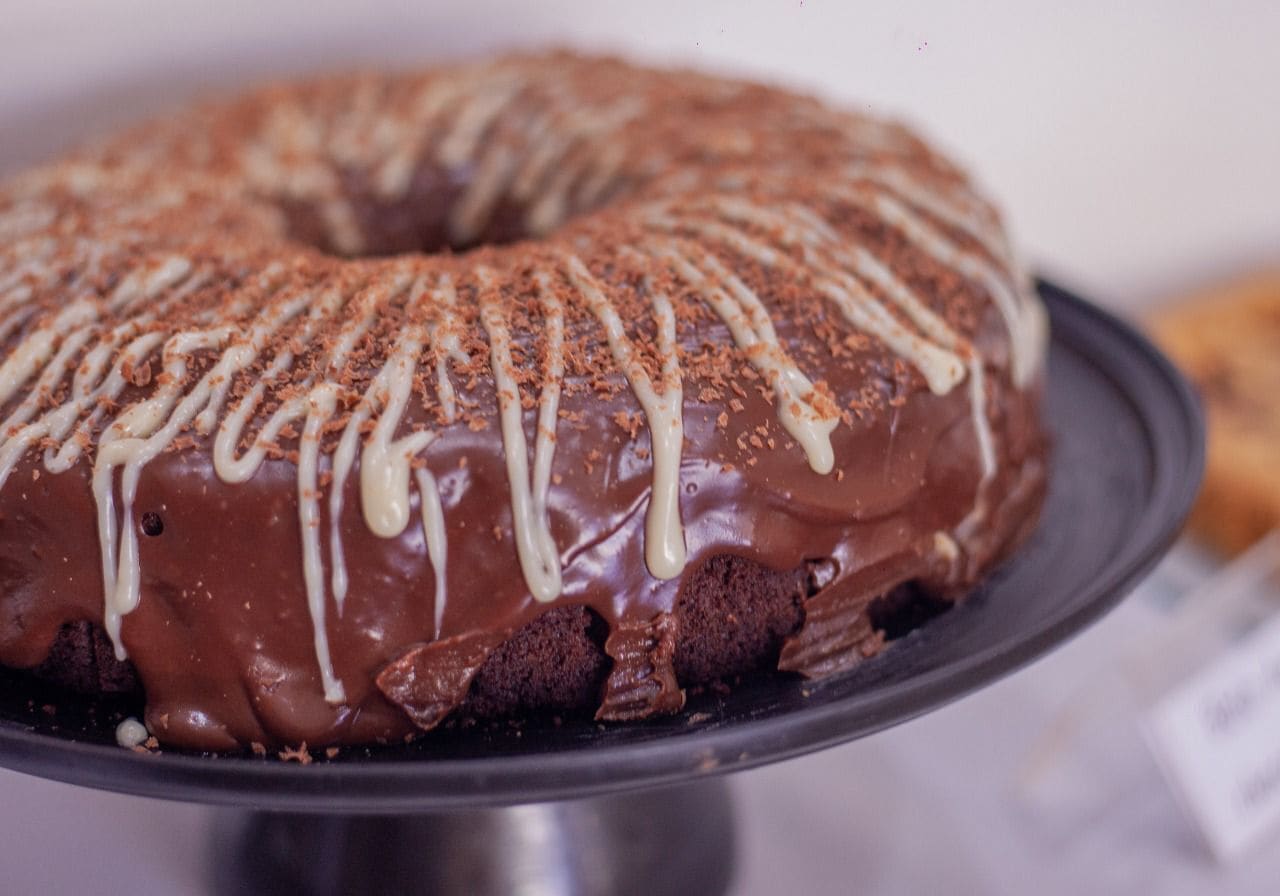 A chocolate cake with white icing and chocolate shavings on a metal stand.