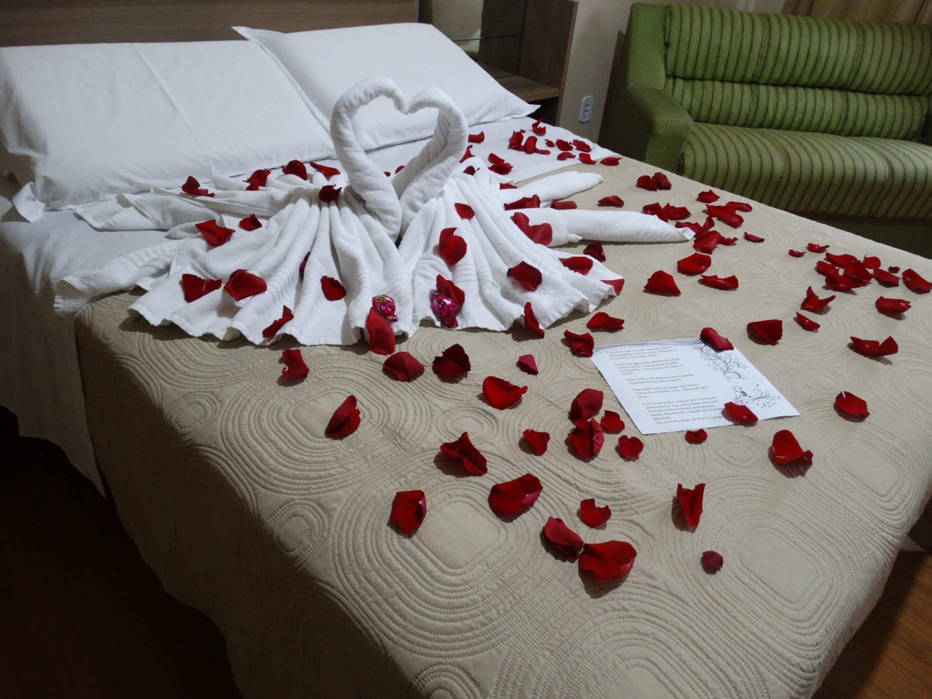 A hotel room with a bed covered in rose petals and a heart-shaped towel arrangement.