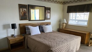 A neatly arranged bedroom with a large bed, a nightstand with a lamp, a bedside table, a mirror, and a window with a view. - Rainbow Inn & Cottages (Carrizozo)