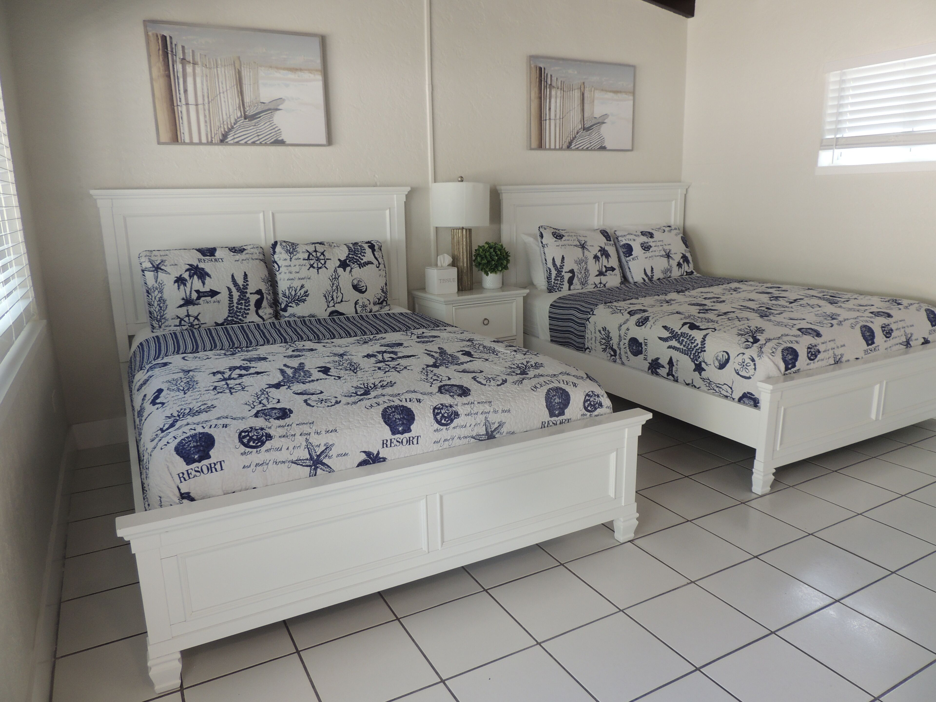 Two white beds with ocean-themed bedding, a white nightstand with a lamp, and framed artwork on the walls.