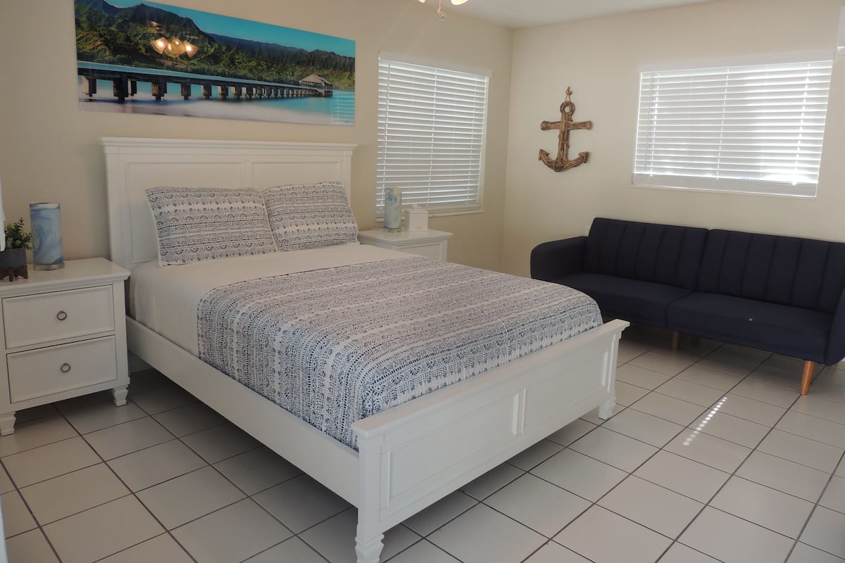 A bedroom with a bed, a nightstand, a dresser, a dark sofa, a ceiling fan, and a painting of a bridge over water.
