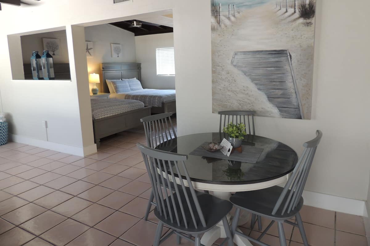 A dining area with a round table and four chairs, a painting of a beach scene, and a bedroom with a bed and a nightstand.