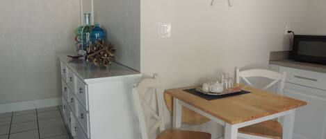 Una piccola cucina pulita con mobili bianchi, un tavolo di legno e due sedie bianche. Si nota una ruota di timone decorativa appesa al muro e una bottiglia blu sul ripiano.