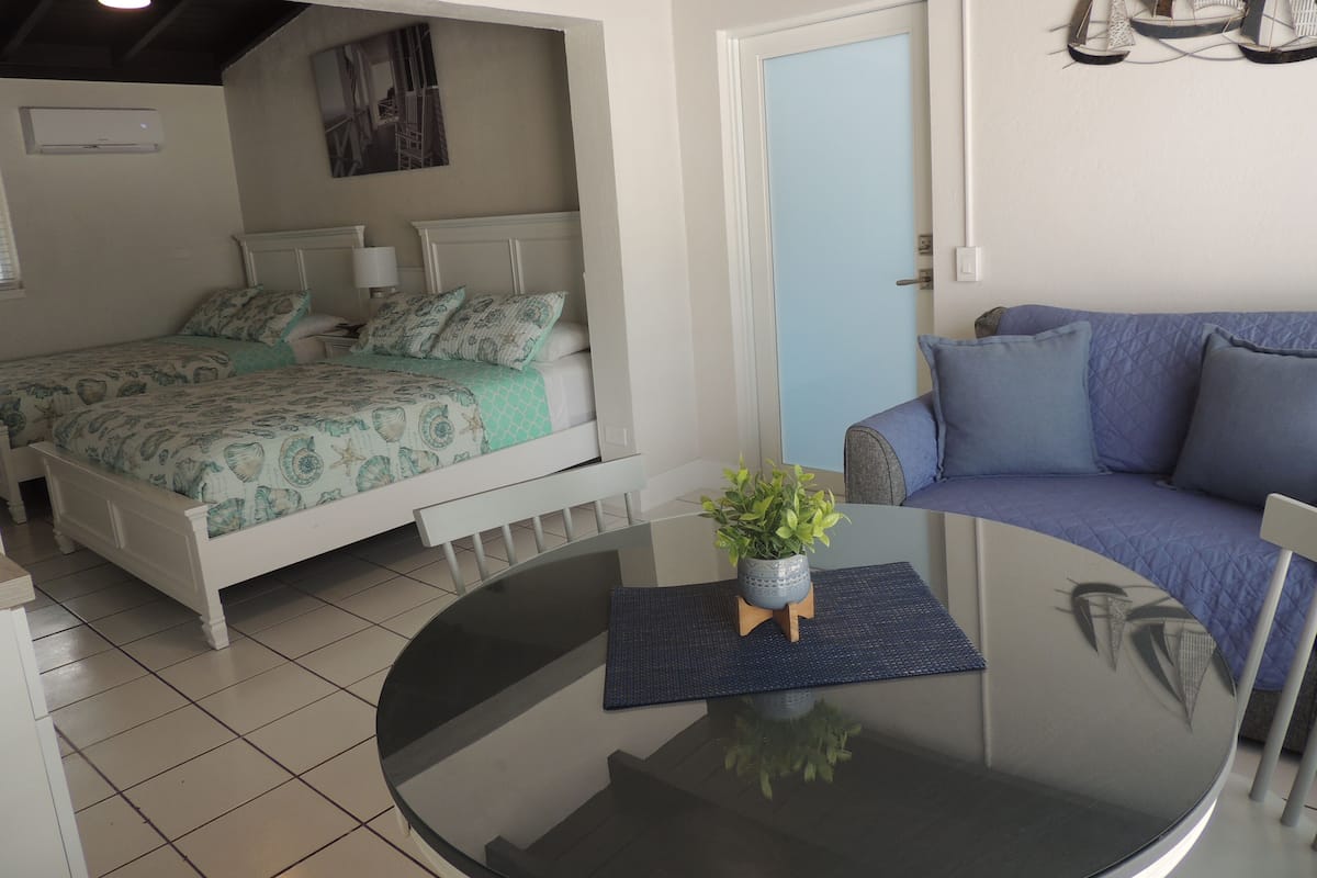 A bedroom with a bed, a sofa, a small table with a plant, and a door with a light switch.