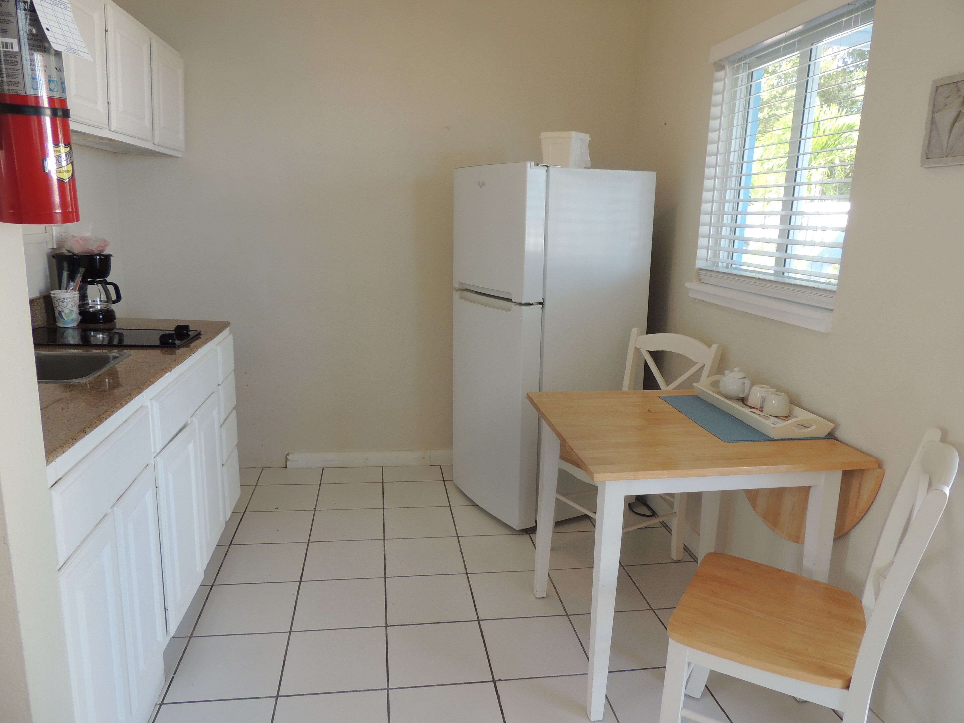 A small kitchen with white cabinets, a white refrigerator, a wooden table, and a window with blinds.