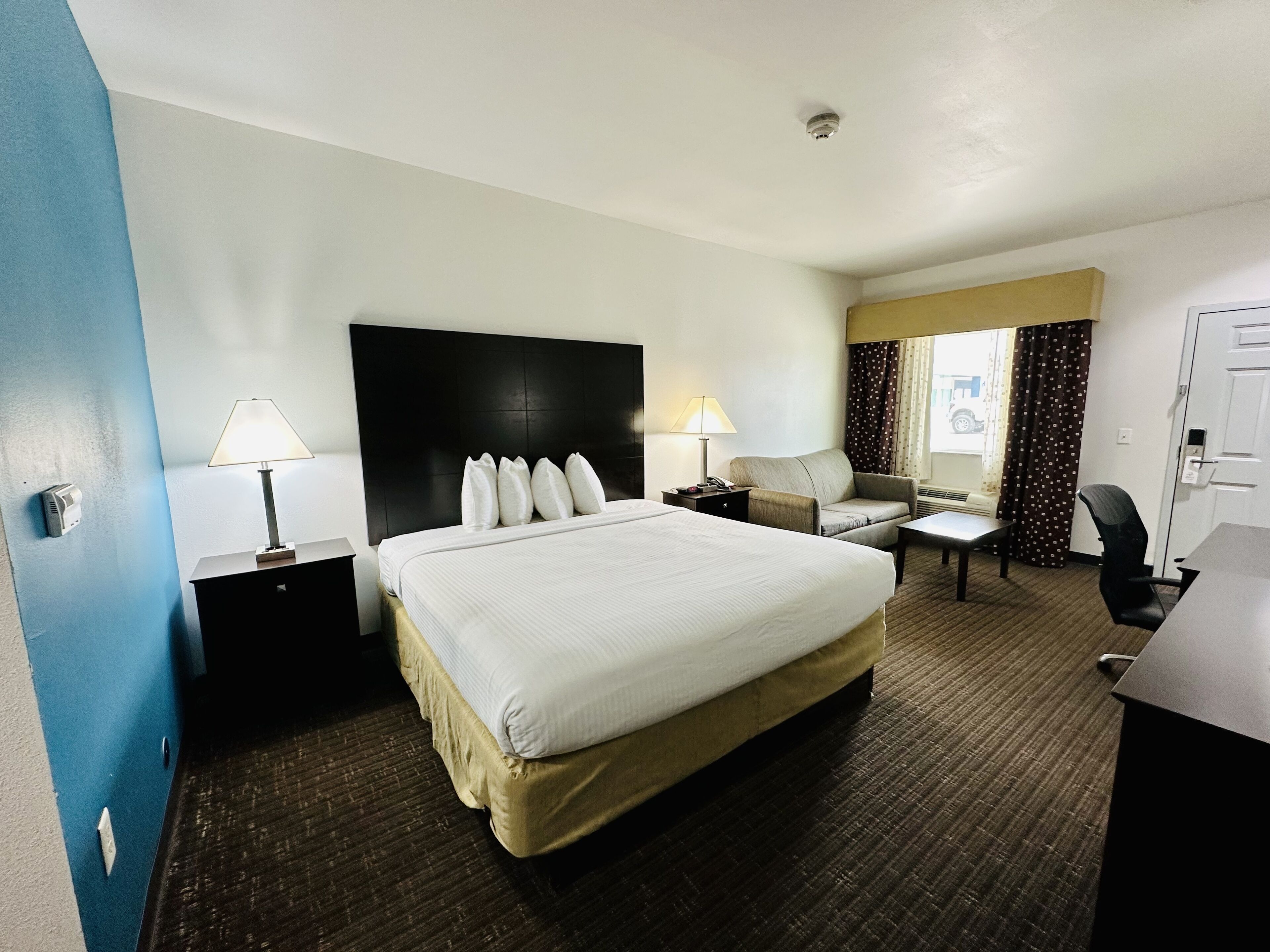 A hotel room with a large bed, two bedside tables with lamps, a desk, a chair, and a window with curtains.