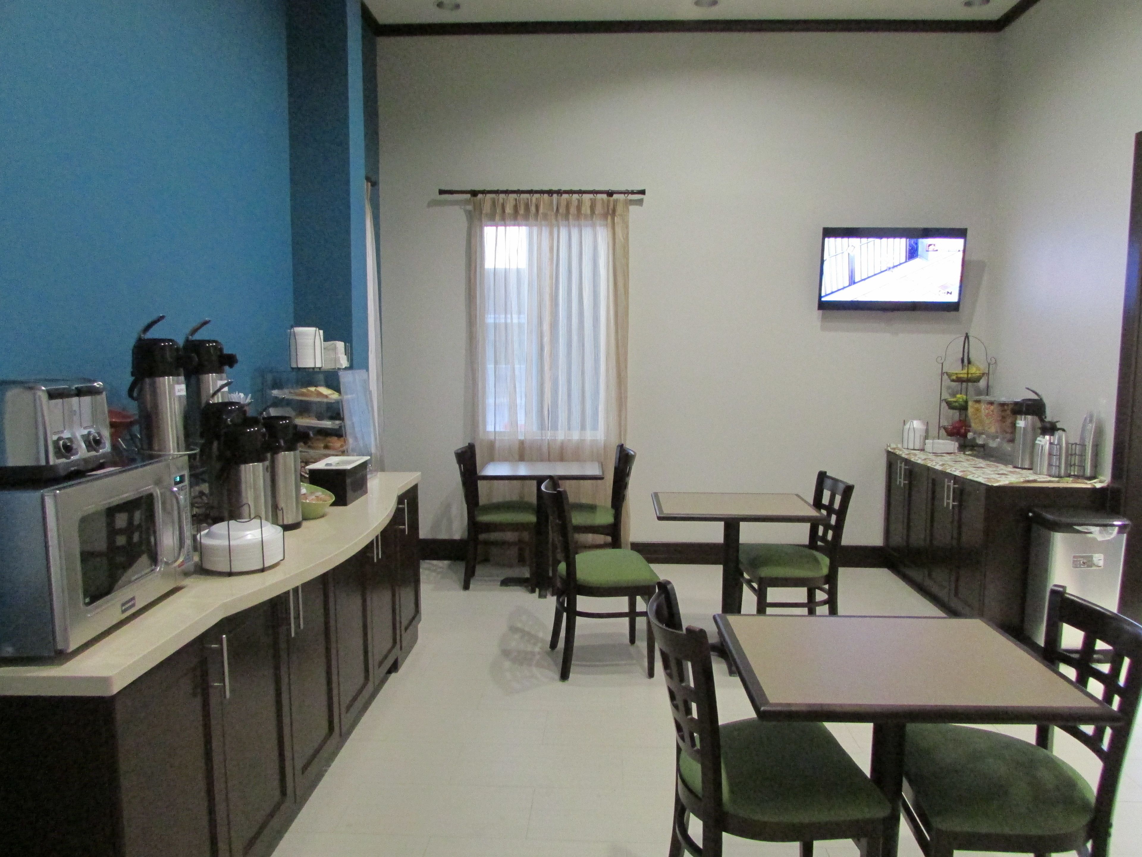A compact kitchen with a microwave, toaster, and coffee maker. There are two dining tables with chairs, a window with curtains, and a television on the wall.