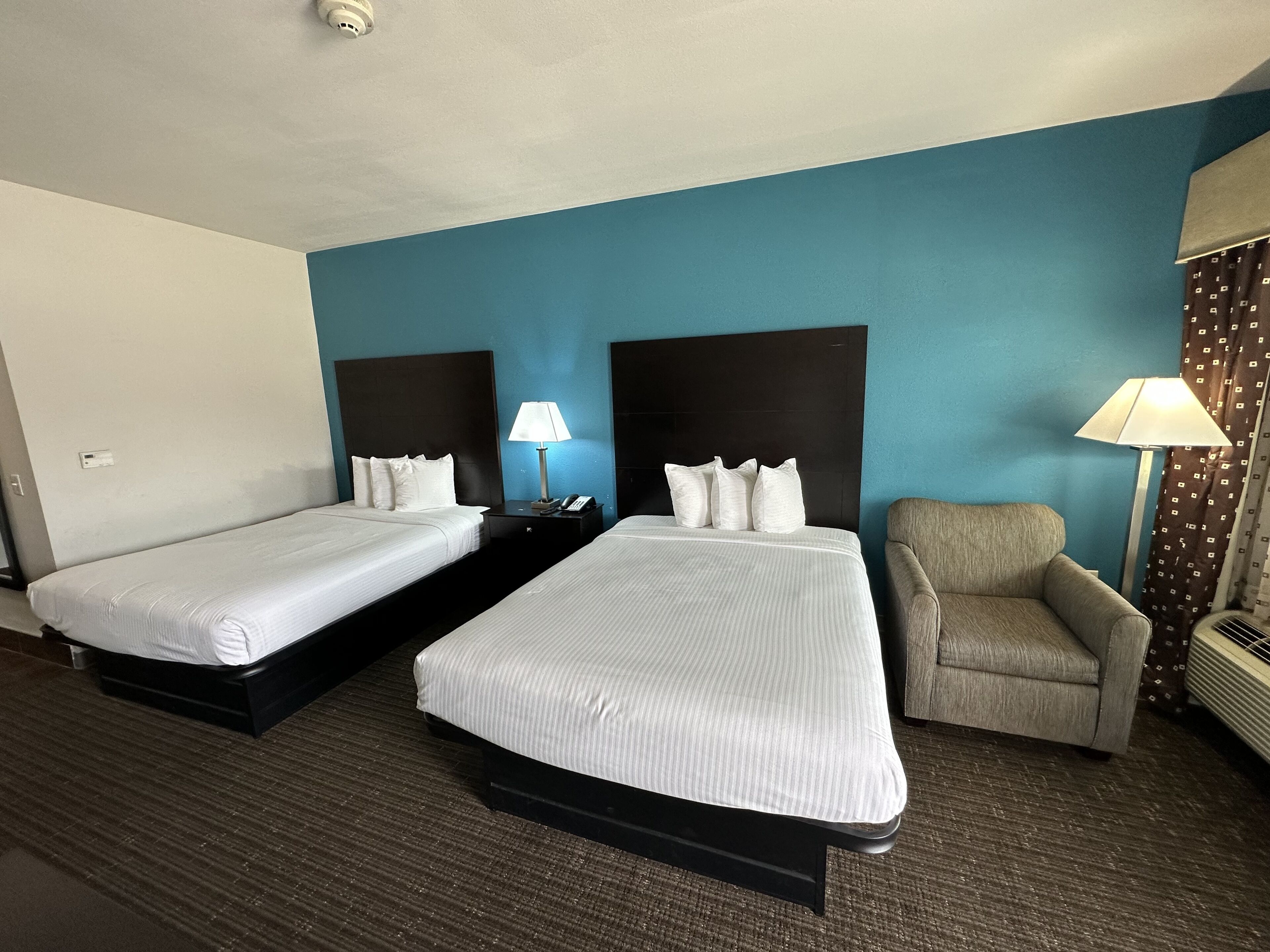 Habitación de hotel con dos camas individuales, una silla, una mesita de noche con lámpara y un calentador.