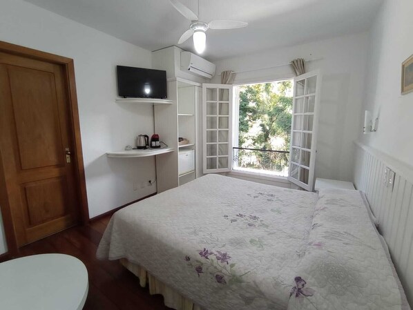 Un dormitorio con cama, televisor sobre una repisa, ventana con cortinas y ventilador de techo.