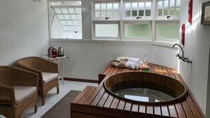 Una habitación con una bañera de madera, sillas de mimbre, una mesita con un hervidor y un ventanal de varios cristales.