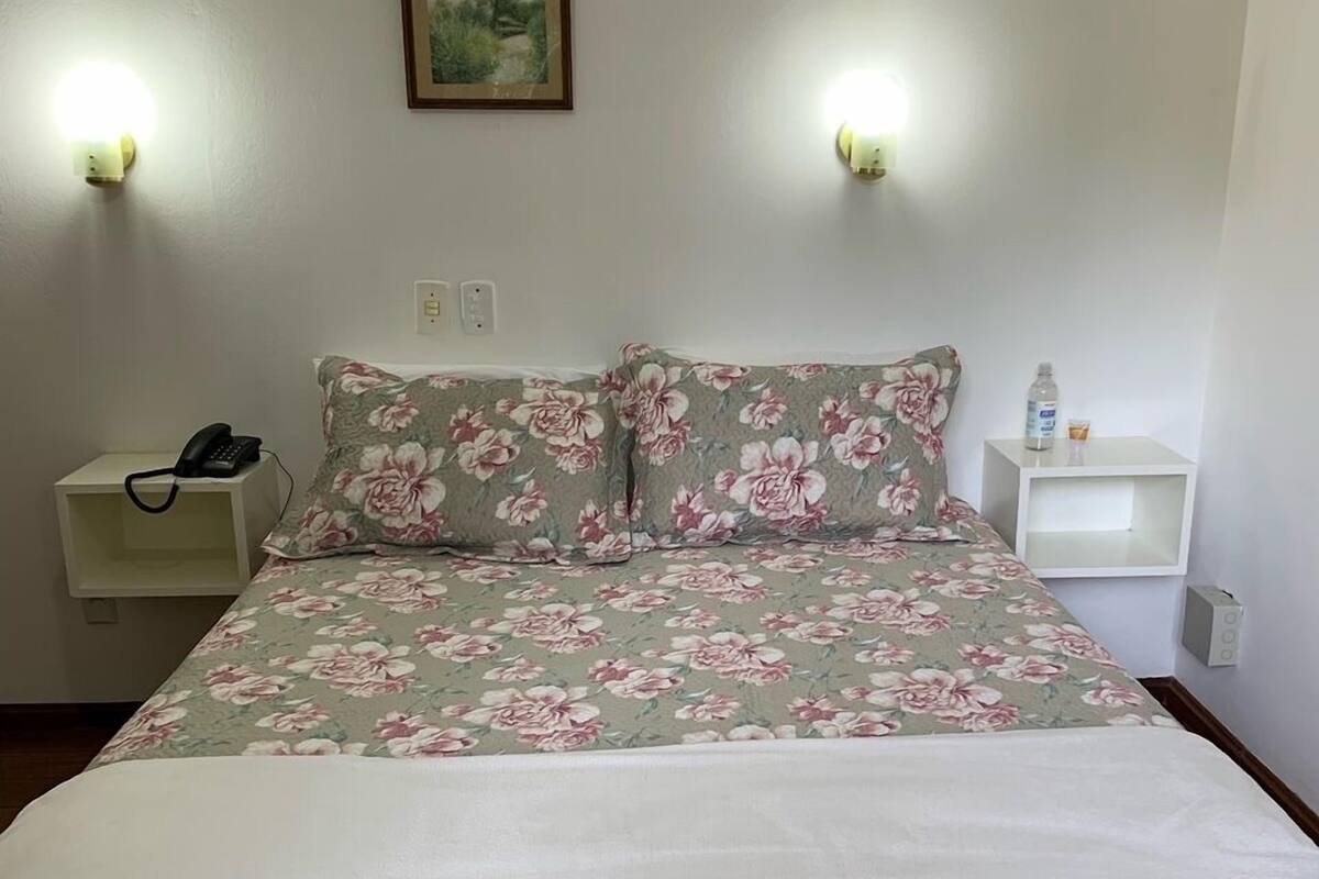 Cama bem arrumada com roupa de cama florida, duas mesas de cabeceira com abajures e um quadro fixado na parede.
