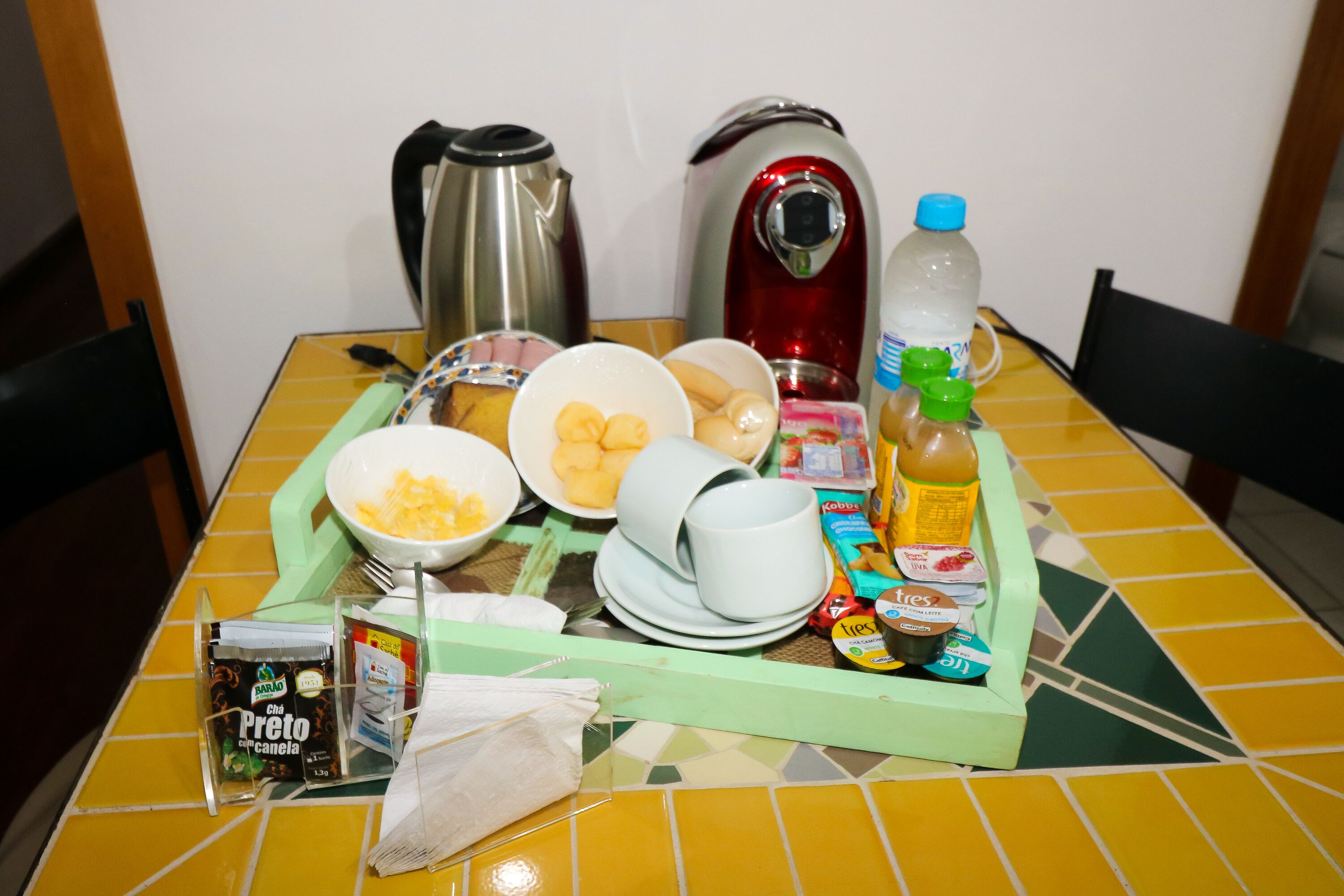 Bandeja de desayuno con alimentos, una botella de agua, una cafetera y un hervidor de té sobre una mesa con azulejos amarillos.
