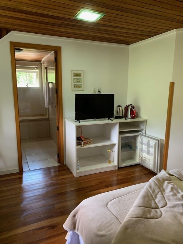 Una habitación con techo de madera, un televisor sobre un soporte blanco, un refrigerador pequeño y una cama con cubrecama blanco.