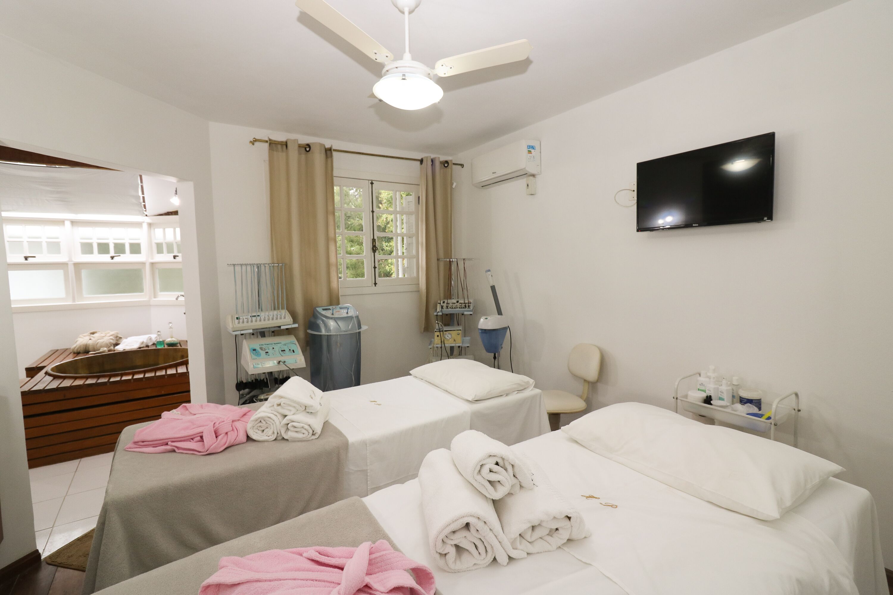 Een hotelkamer met twee bedden, een televisie, een raam met gordijnen, een plafondventilator en een badkamer met een wastafel en spiegel.