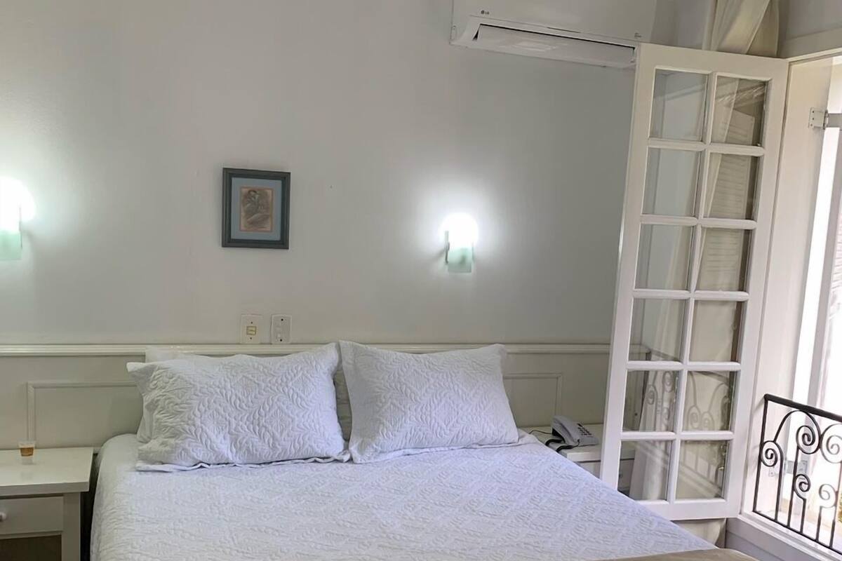 Quarto com cama, mesa de cabeceira, ar condicionado fixado na parede e janela com grade de metal.