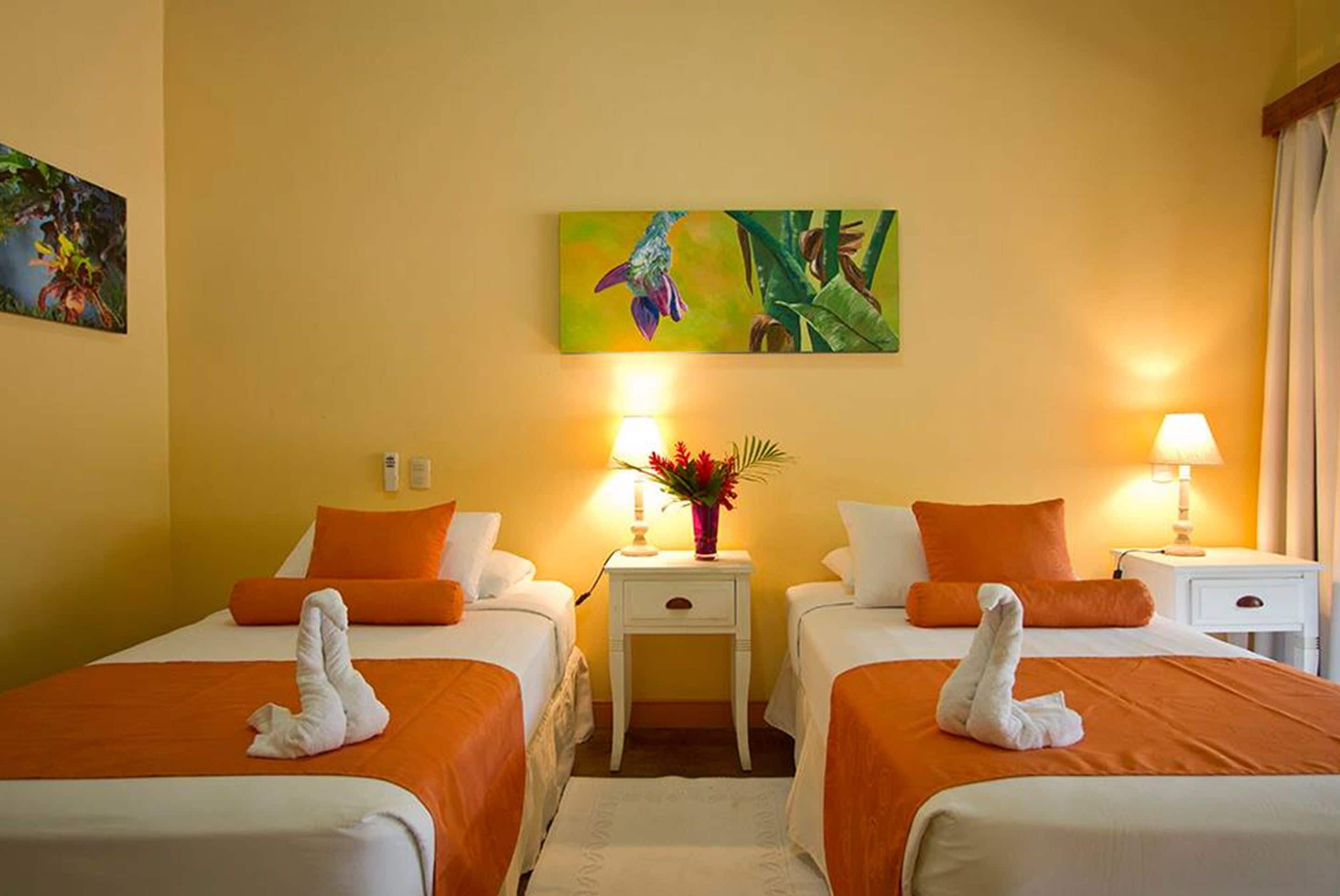 A hotel room with two beds, each with an orange bedspread and white pillows, a small white nightstand with a lamp and a vase of flowers, and a painting of a bird on the wall.