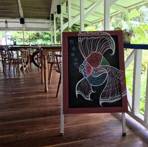 A wooden deck with tables and chairs, a framed artwork of a fish, and a view of greenery through a glass railing. - Villas Del Caribe (Cahuita)