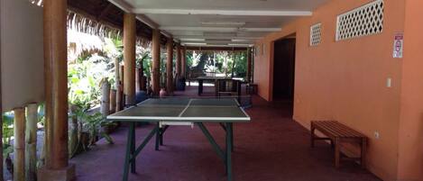 Un espacio al aire libre techado con mesa de ping pong, bancos y una estructura de techo de paja.