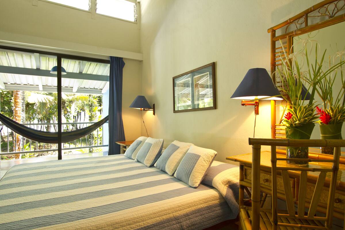 A bed with striped bedding, a hammock, a bedside table with a lamp, and a view of palm trees through large windows.