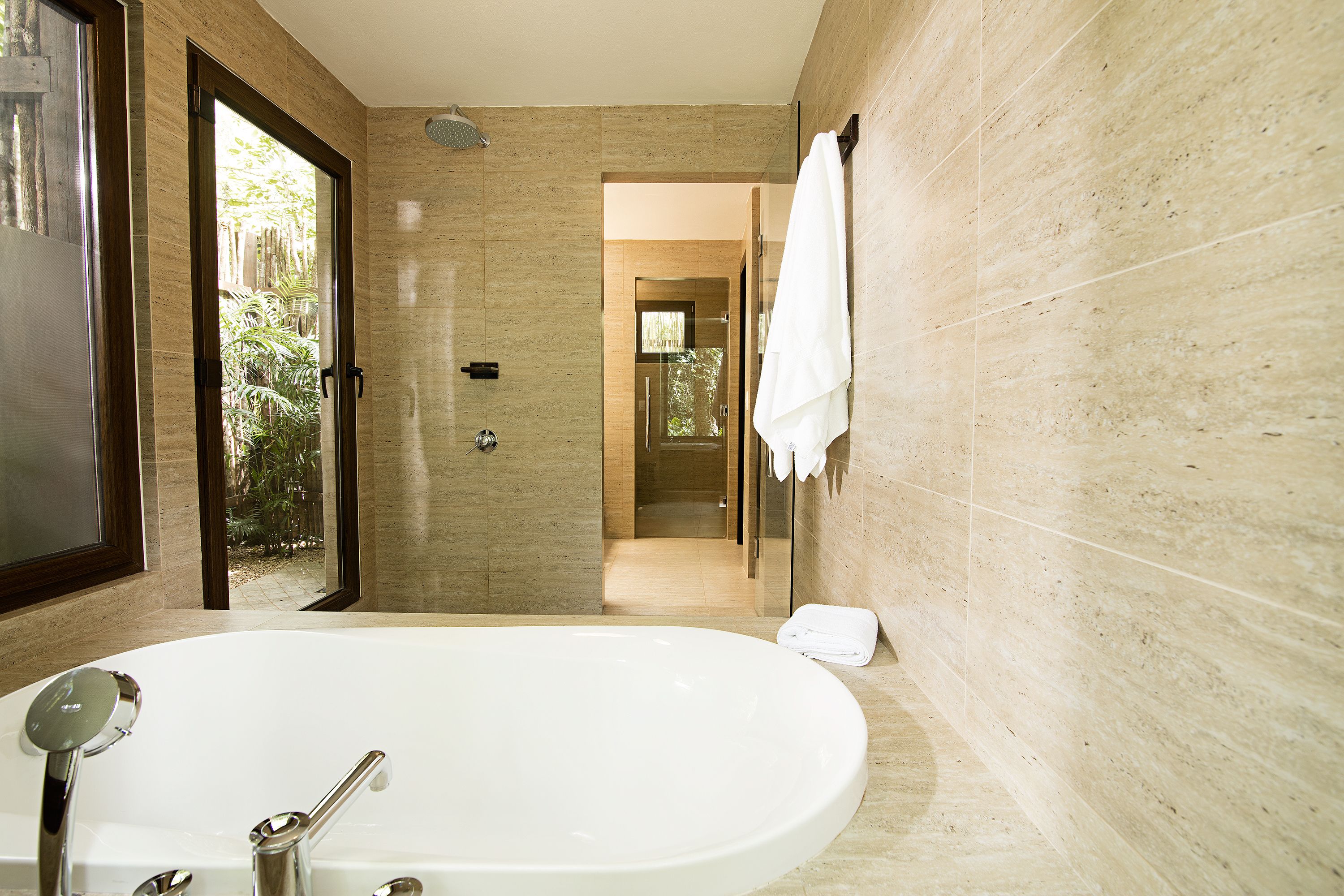 Une salle de bain moderne dotée d’une grande baignoire, d’une douche à parois de verre et offrant une vue sur un jardin grâce à une porte ouverte.