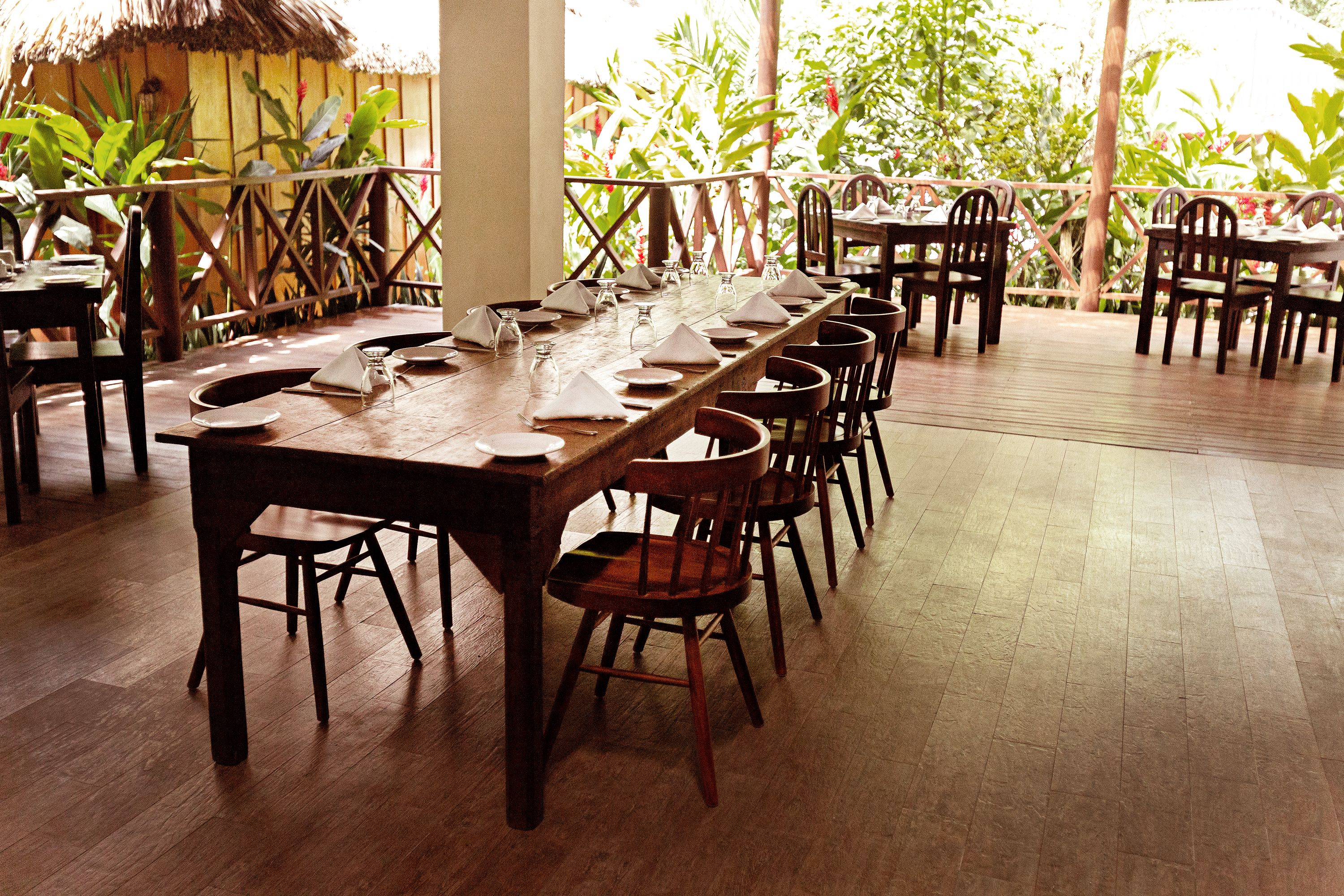 A long wooden dining table set with chairs, surrounded by a wooden deck with a railing, and lush greenery in the background.