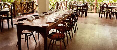 A long wooden dining table set with chairs, surrounded by a wooden deck with a railing, and lush greenery in the background.
