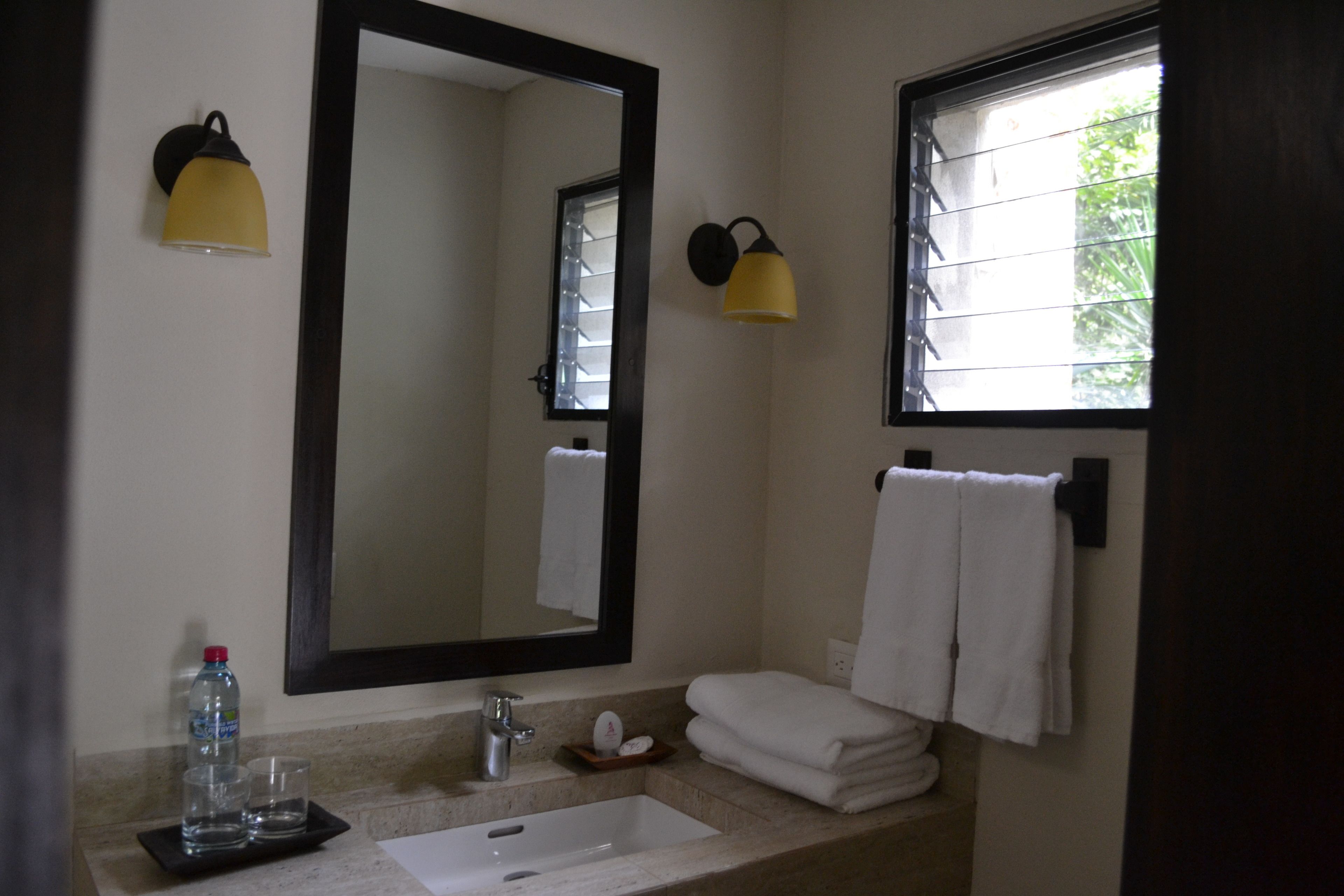 Une salle de bain dotée d’un grand miroir, de deux lampes fixées au mur, d’un lavabo, d’une fenêtre offrant une belle vue et d’un porte-serviettes avec des serviettes pliées.
