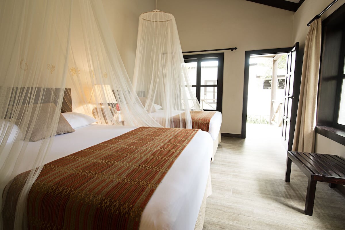 A hotel room with two beds, a wooden bench, and a large window.