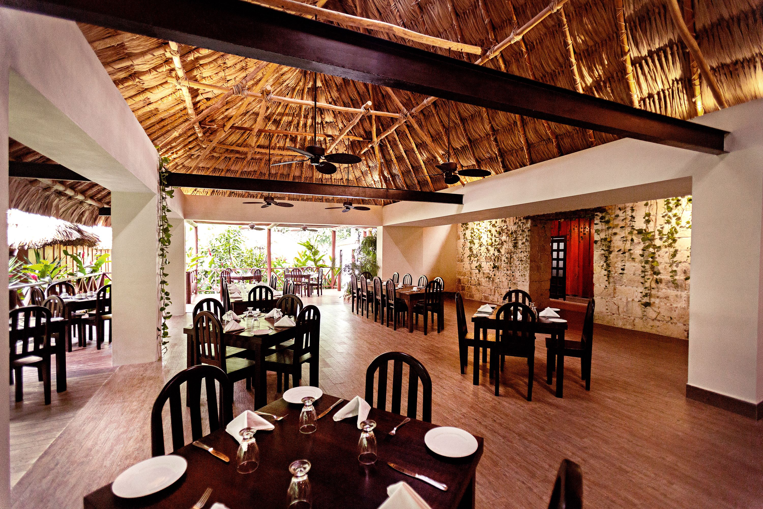 A restaurant with wooden tables and chairs, a thatched roof, and a stone wall.