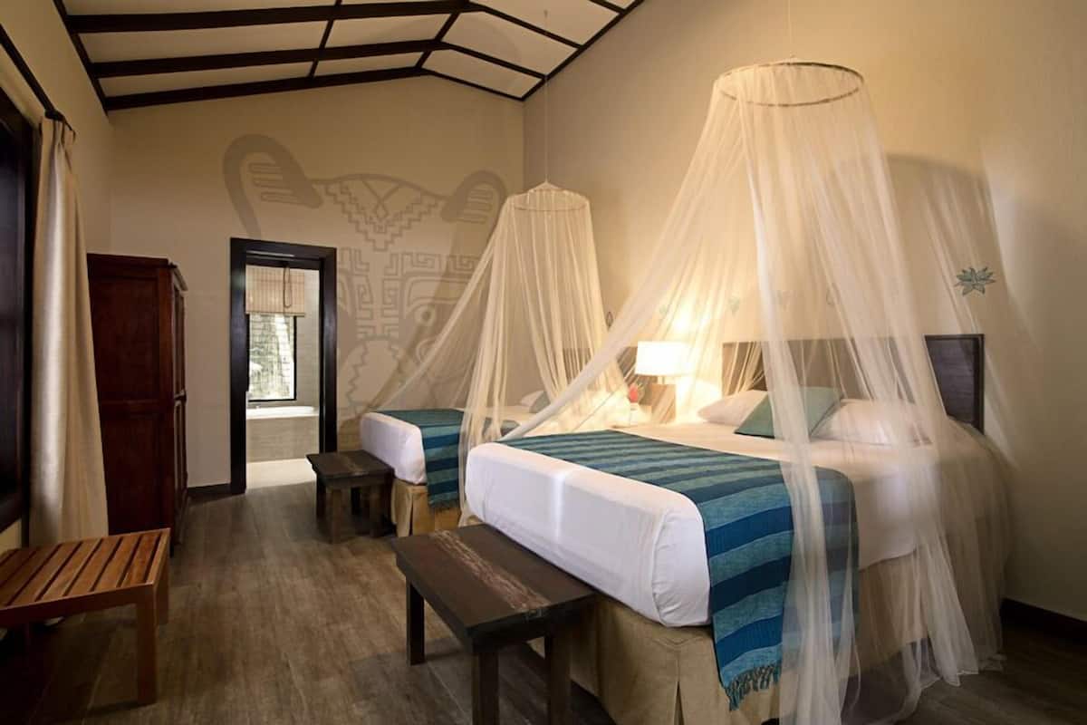 A room with two beds, each with a canopy, a bench, and a wooden table.