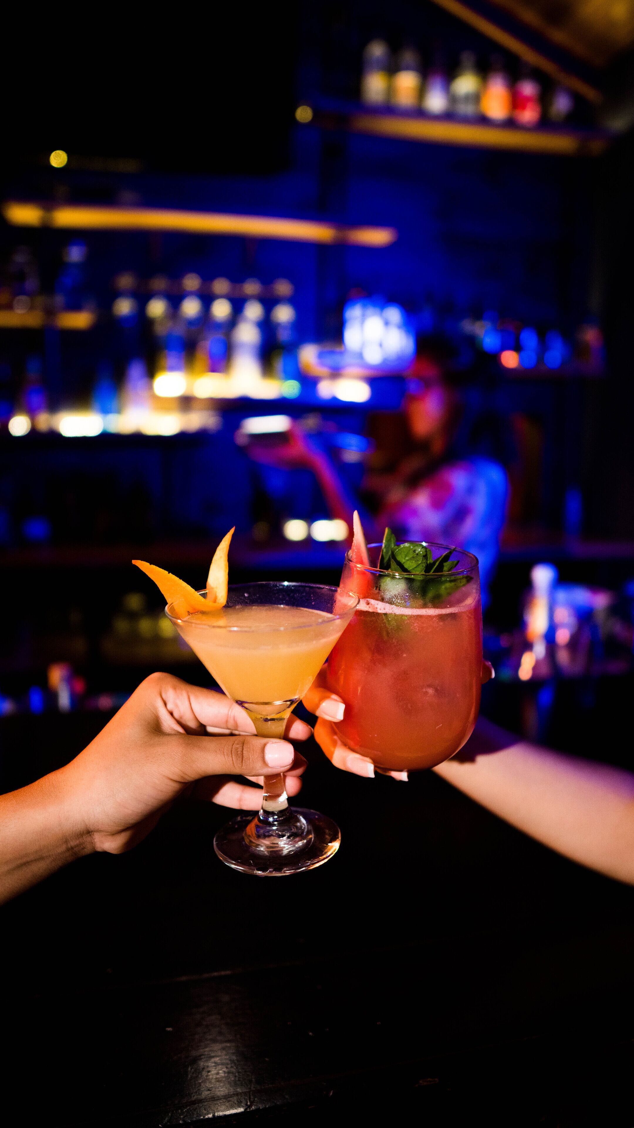 Dos cócteles sostenidos por manos en un bar.