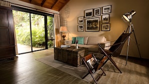A living room with a wooden floor, a sofa, a coffee table, a lamp, and a telescope. There are framed pictures on the wall and a window with curtains. - Jungle Lodge Hotel (Flores)