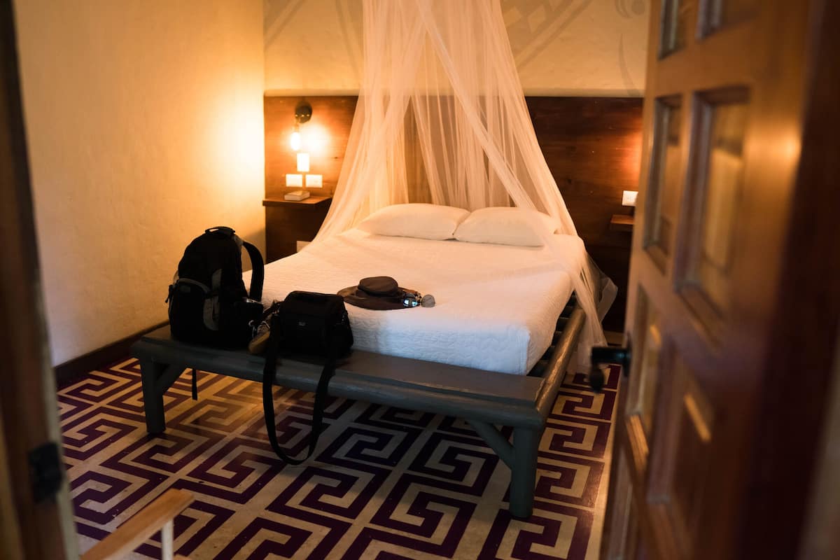 A hotel room with a bed, a bench, a suitcase, and a hat.