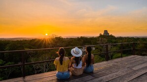 Three people sitting on a wooden deck overlooking a sunset with a view of a distant ancient structure. - Hostal Jungle Lodge Tikal (Flores)
