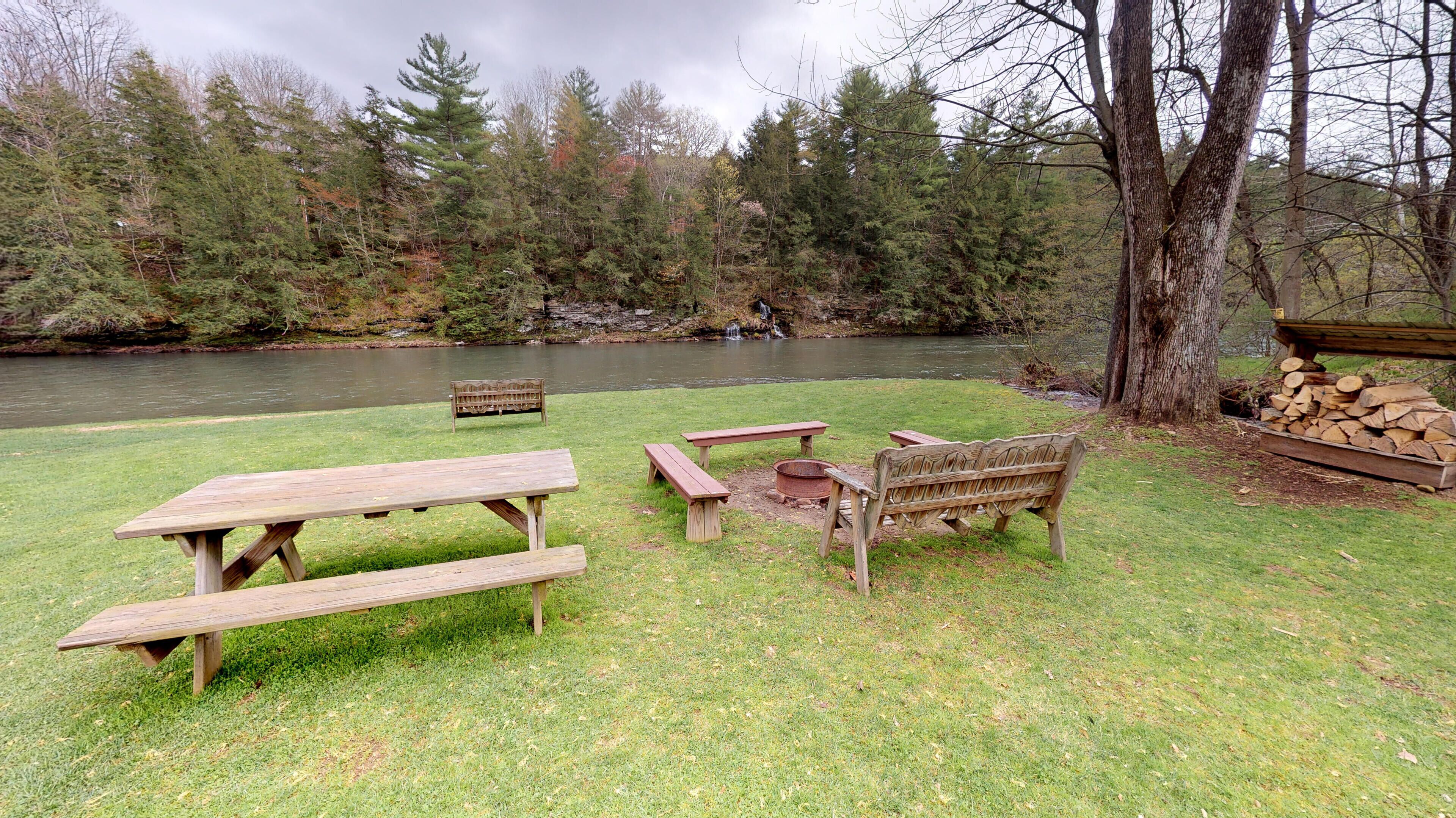 A picnic area with wooden benches and picnic tables by a river, surrounded by trees.