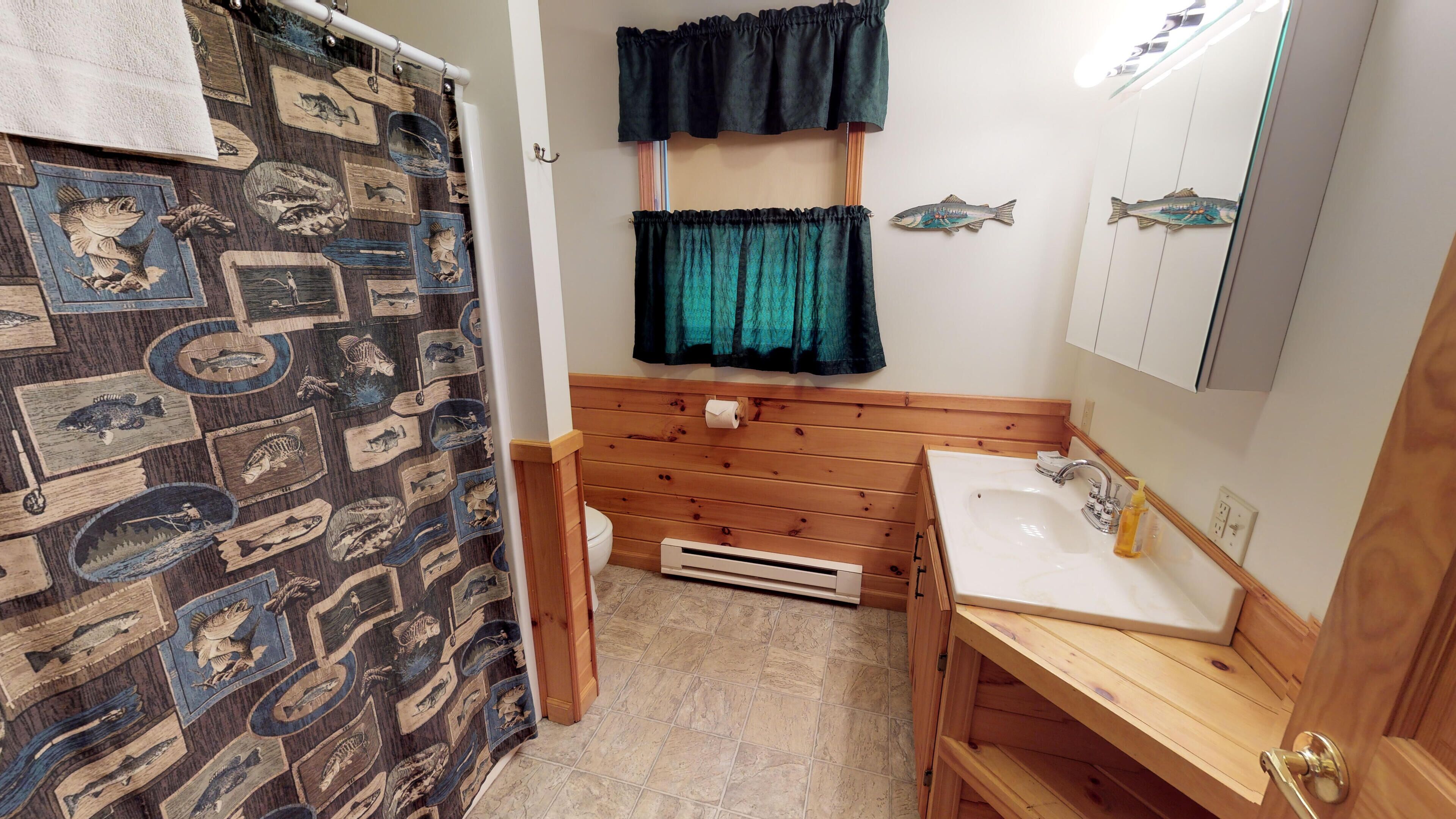 Une salle de bain avec un meuble-lavabo en bois, un évier et un miroir. On y trouve un rideau de douche orné d’un motif de poissons, et les toilettes sont visibles.