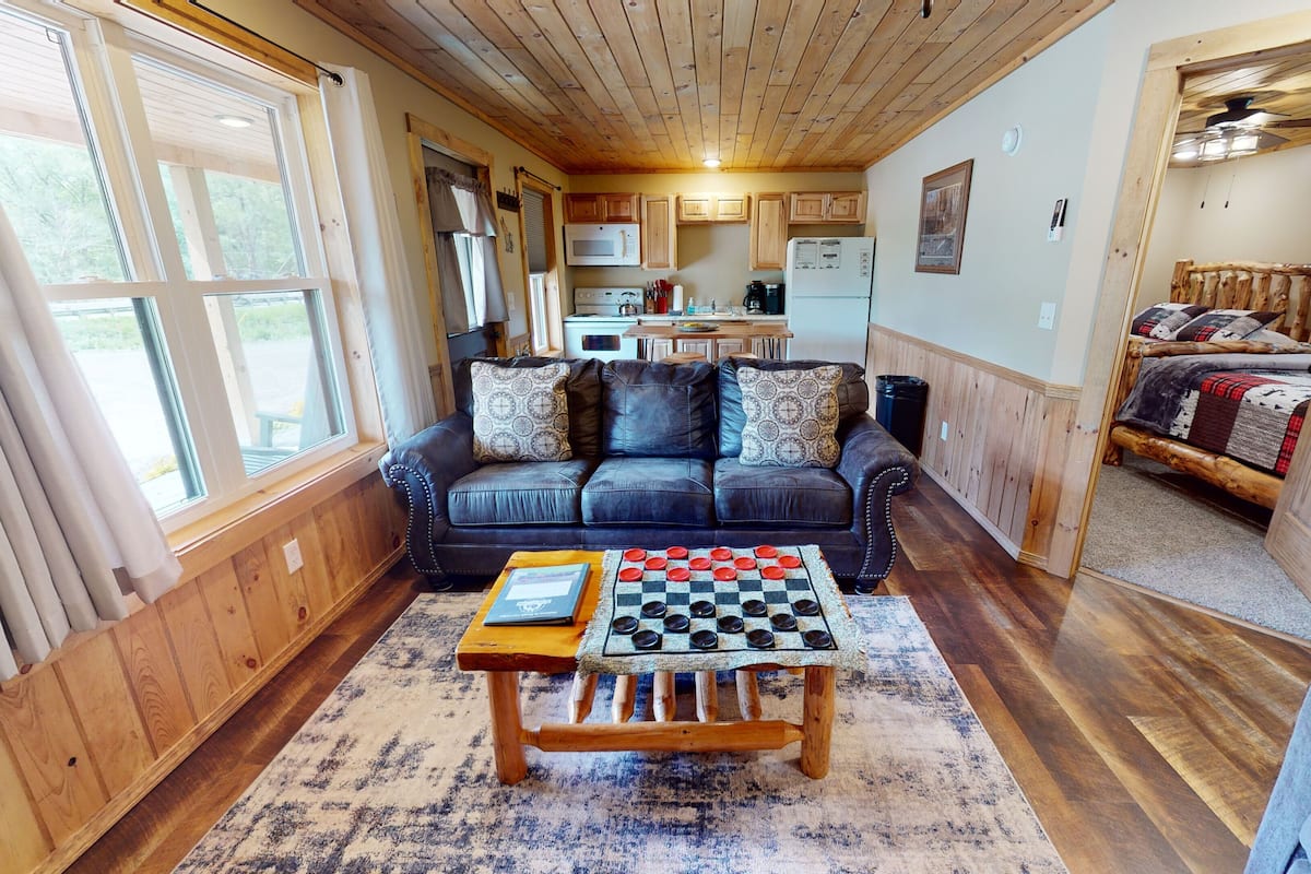 A cozy living room with a wooden ceiling, a black leather sofa, a checkered coffee table, and a view of a kitchen and bedroom through an open door.