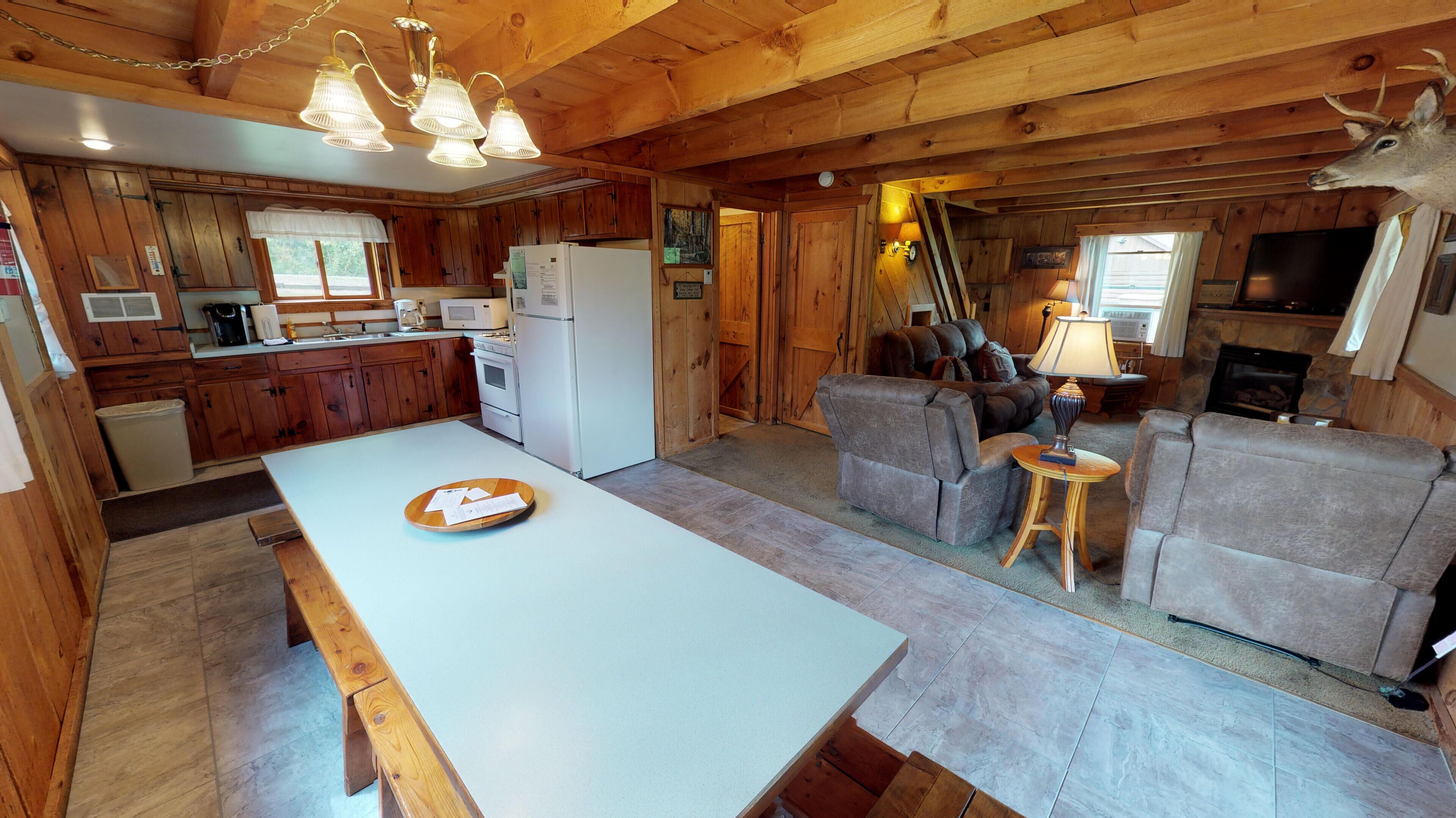 A cabin interior with a wooden dining table, kitchen area, and a living room with a fireplace.