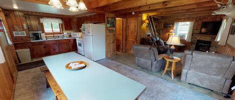 A cabin interior with a wooden dining table, kitchen area, and a living room with a fireplace.